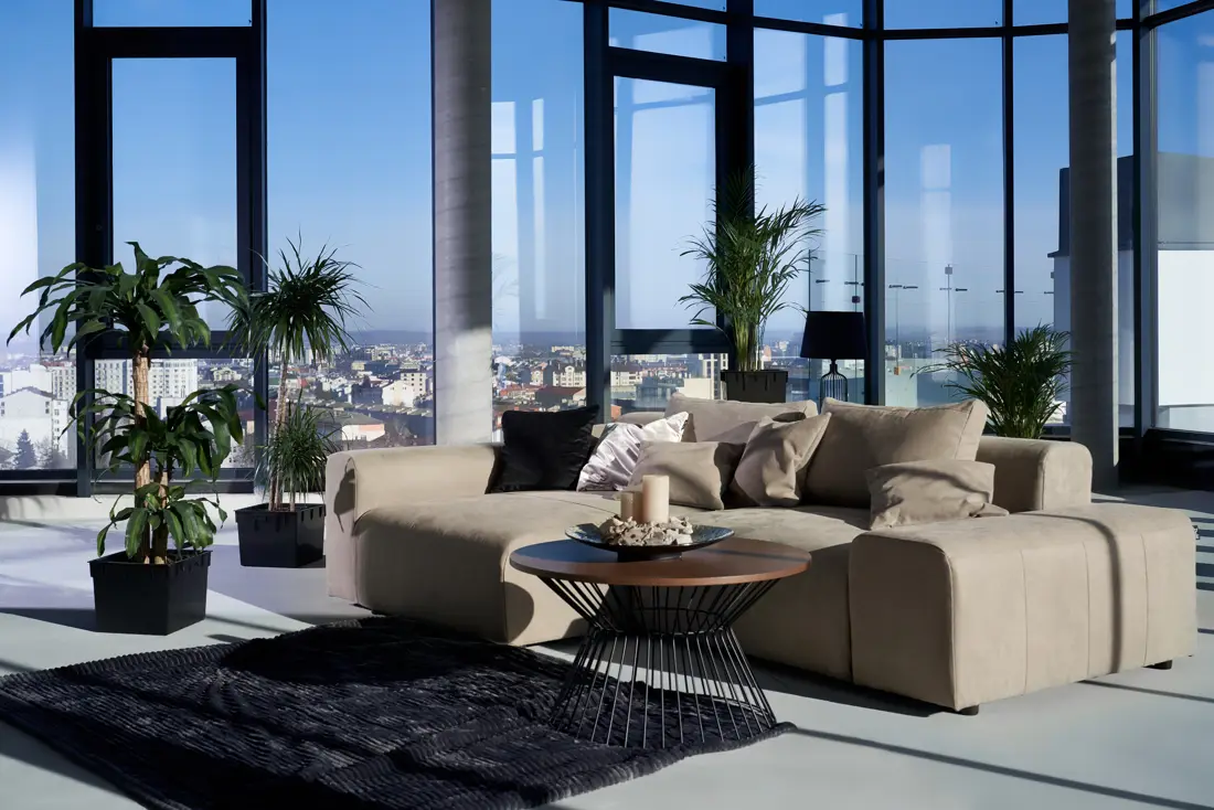 Sala do apartamento decorado com sofá claro, plantas e janelas do piso ao teto.
